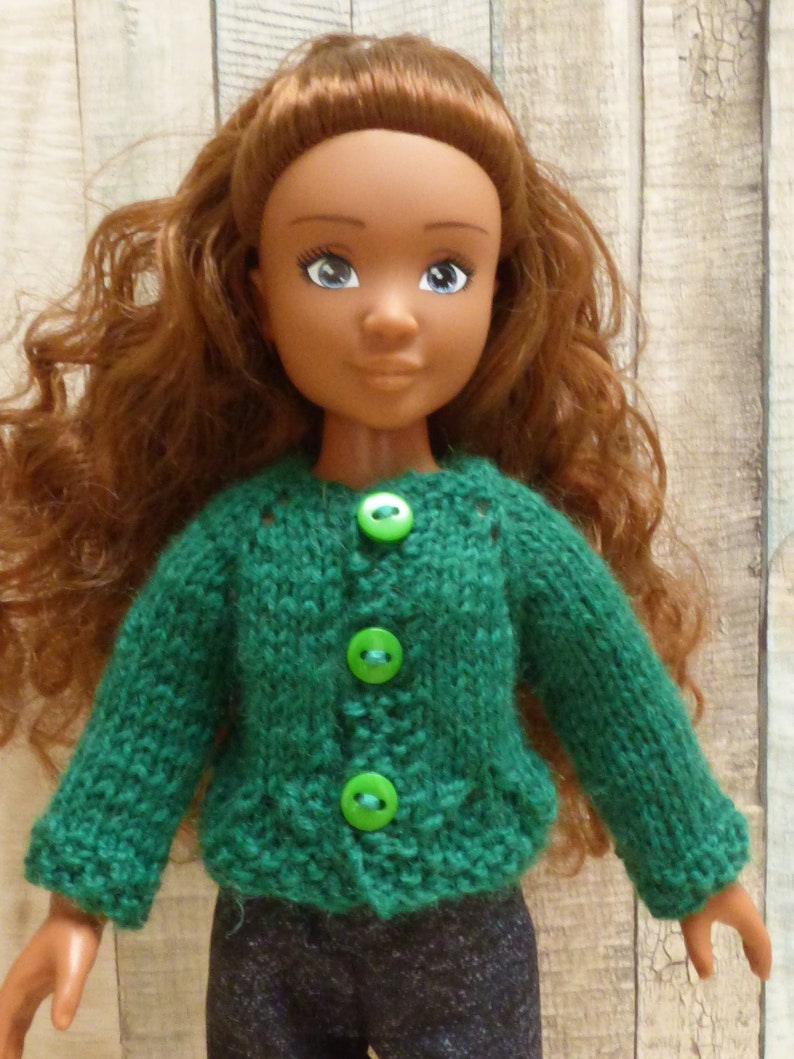 May include: A doll with long brown curly hair wearing a green knitted cardigan with green buttons. The doll is wearing dark blue jeans.