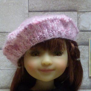 Peut inclure: Un béret rose en tricot avec une texture duveteuse. Le béret est porté par une poupée aux cheveux bruns et aux yeux bleus.