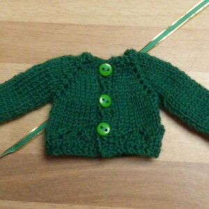 May include: A green knitted cardigan with four green buttons. The cardigan is made with a simple knit stitch and has a classic design.