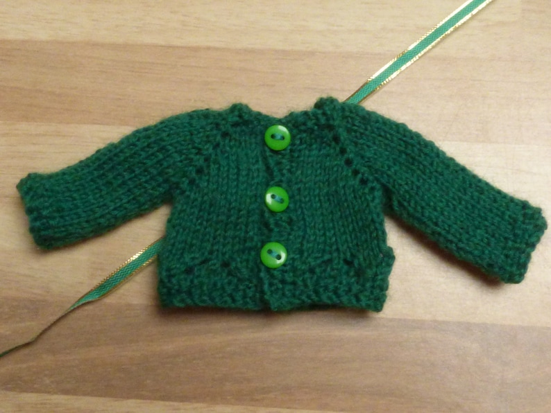 May include: A green knitted cardigan with four green buttons. The cardigan is made with a simple knit stitch and has a classic design.