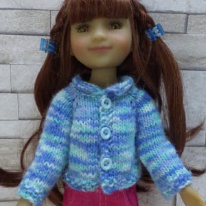 Peut inclure: Une poupée portant un cardigan en tricot rayé bleu et blanc avec des boutons bleus et un pantalon rose.