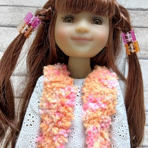 Może przedstawiać: Lalka z brązowymi włosami upiętymi w kucyki, ozdobiona różowymi i pomarańczowymi spinkami do włosów. Lalka ma na sobie białą koszulę z dziurkami, puszystą pomarańczowo-różową kamizelkę i niebieskie dżinsy. Oczy lalki są otwarte.