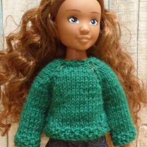May include: A doll with long, curly brown hair wearing a green knitted sweater and blue jeans.