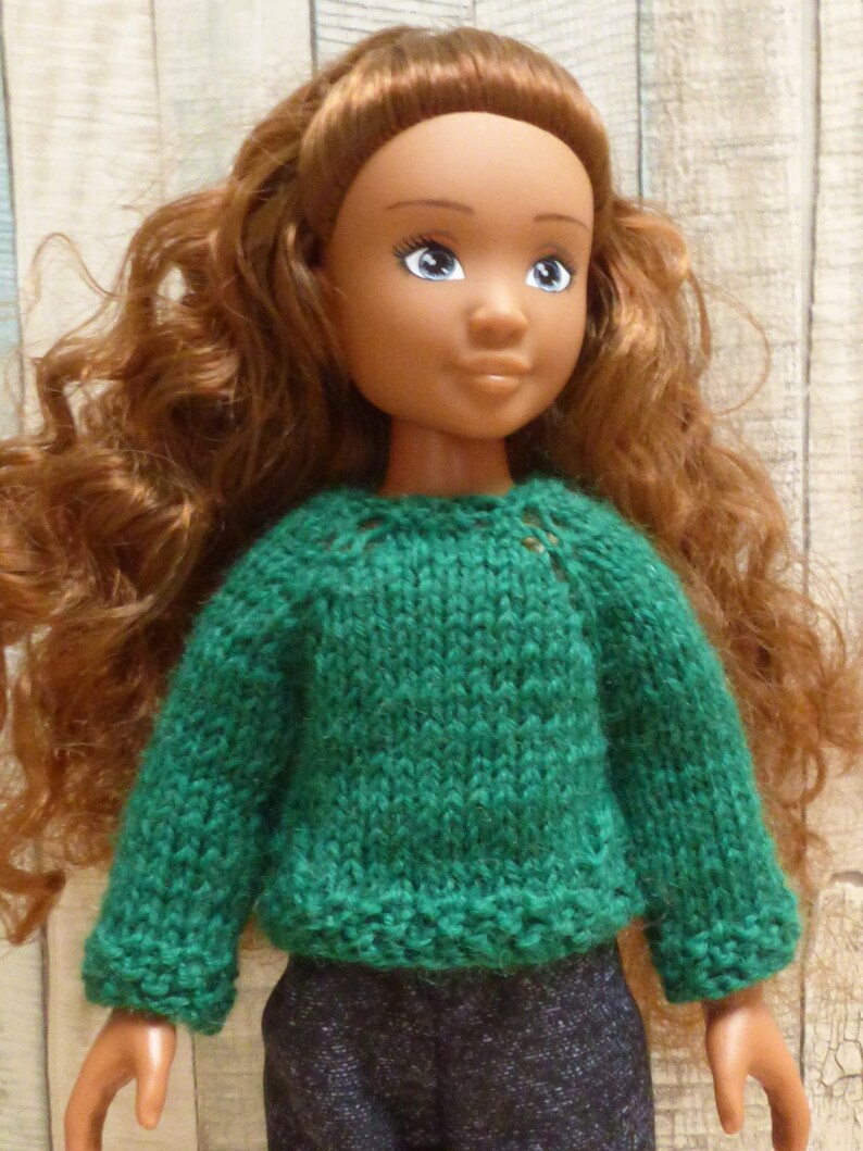 May include: A doll with long, curly brown hair wearing a green knitted sweater and blue jeans.