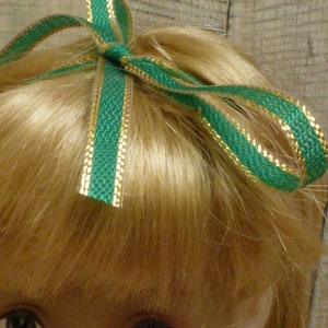 May include: A doll with blonde hair wearing a green and gold ribbon bow.