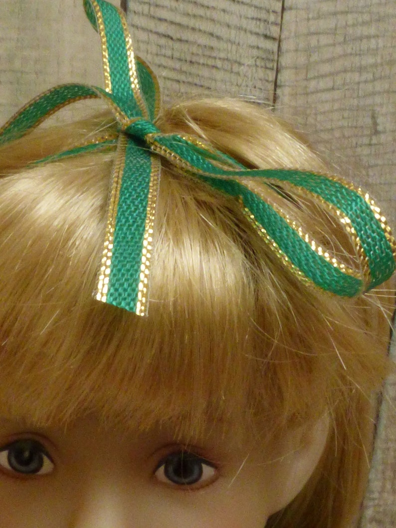 May include: A doll with blonde hair wearing a green and gold ribbon bow.