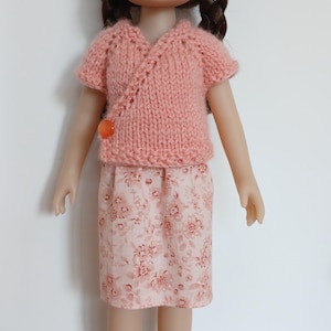 Peut inclure: Une poupée aux cheveux bruns tressés, vêtue d'un haut cache-cœur en tricot pêche à manches courtes avec un bouton orange, assorti d'une jupe rose clair à motif floral et de chaussures noires scintillantes à brides croisées.