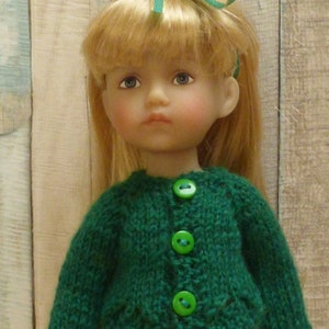 May include: A doll wearing a green knitted cardigan with green buttons. The doll has blonde hair and blue eyes.