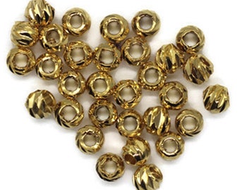 May include: A close-up of a heart-shaped pile of gold-colored beads. The beads are round and have a textured surface. The beads are arranged in a heart shape on a white background.