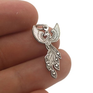 May include: A silver-toned phoenix charm pendant with intricate details. The phoenix is depicted with outstretched wings and a long, flowing tail.