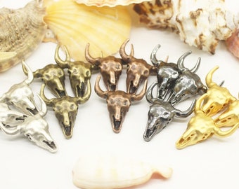 May include: A collection of ten metal bull skull charms in various finishes, including silver, gold, and bronze. The charms are detailed and have a rustic look.