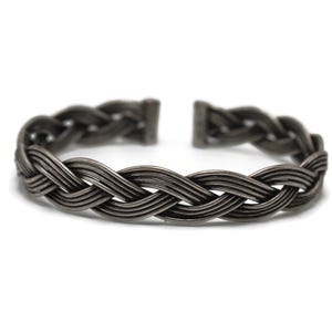 Puede incluir: Una pulsera de puño trenzada de color gris oscuro. La pulsera está hecha de múltiples hebras de metal tejidas juntas en un patrón texturizado. El diseño de puño abierto permite un uso fácil. La pulsera está sobre un fondo blanco.