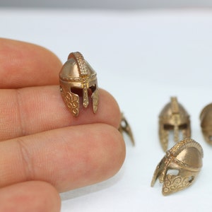 May include: Four small gold-colored metal helmets with intricate details. The helmets are in the shape of a traditional Greek or Roman warrior helmet.