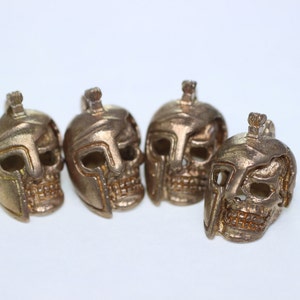 May include: Four gold-colored skull charms with a helmet design. The skulls are detailed with eye sockets, a nose, and a mouth.