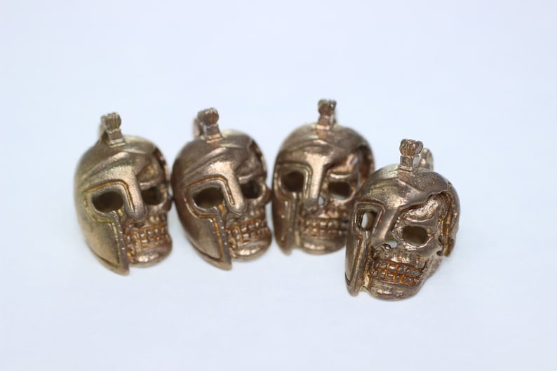 May include: Four gold-colored skull charms with a helmet design. The skulls are detailed with eye sockets, a nose, and a mouth.
