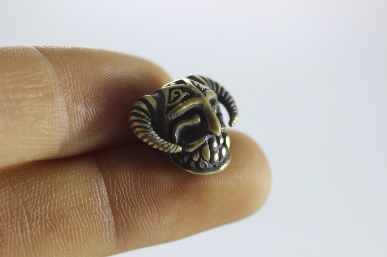 May include: A small, detailed, brass charm in the shape of a horned, mythical creature's head. The charm is a dark bronze color with gold accents.