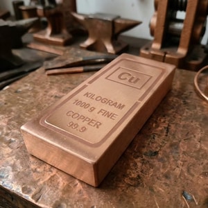 May include: A copper ingot, stamped with "Cu" and details: "KILOGRAM 1000 g FINE COPPER 99.9". The rectangular bar is displayed on a copper-colored surface, with tools and copper coils in the background.