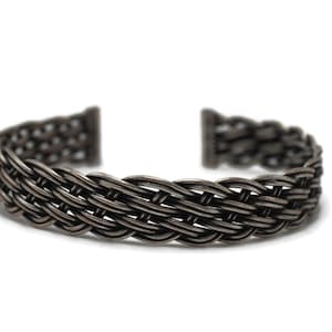 Peut inclure: Un bracelet manchette en métal gris foncé avec un motif tressé. Le bracelet a un aspect texturé et est ouvert aux extrémités. Le fond est blanc uni.
