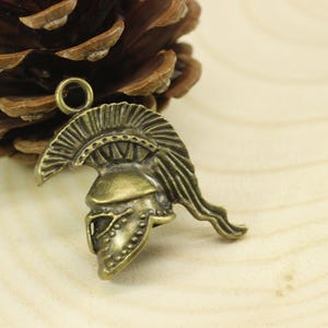 May include: A bronze-colored metal charm in the shape of a Roman soldier's helmet. The helmet has a detailed design with a plume on top and a face mask.
