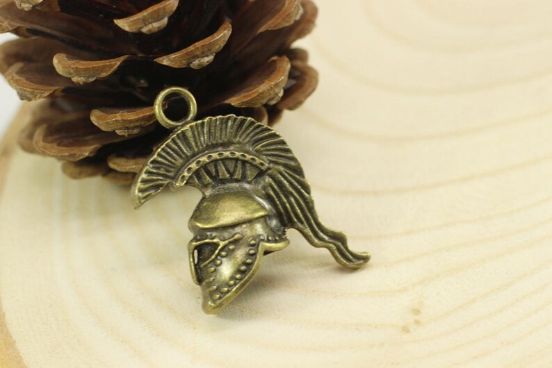 May include: A bronze-colored metal charm in the shape of a Roman soldier's helmet. The helmet has a detailed design with a plume on top and a face mask.