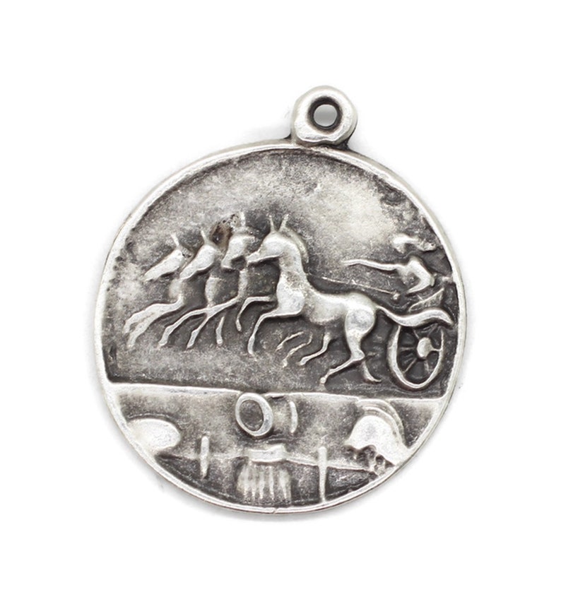 May include: Silver charm featuring a detailed relief of a chariot pulled by four horses. The chariot driver is visible in the background. The charm is round and has a loop at the top for attaching to a necklace or bracelet.