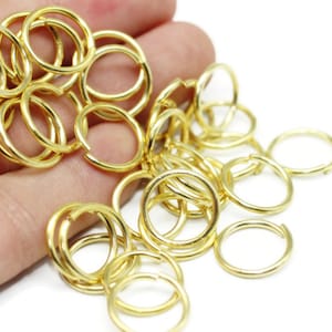 May include: A collection of small, gold-colored jump rings. These open rings are used for jewelry making and other crafts. The rings are round and have a bright, shiny finish. They are scattered on a white surface.