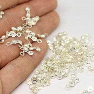Peut inclure: Un tas de connecteurs métalliques argentés avec deux boucles sur chaque pièce. Les pièces sont éparpillées sur une surface blanche.