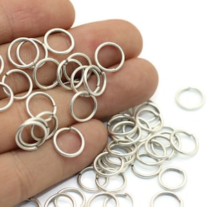 May include: A large quantity of silver jump rings, a type of jewelry finding, are shown in a hand. The jump rings are small and circular, and they are used to connect different pieces of jewelry together.