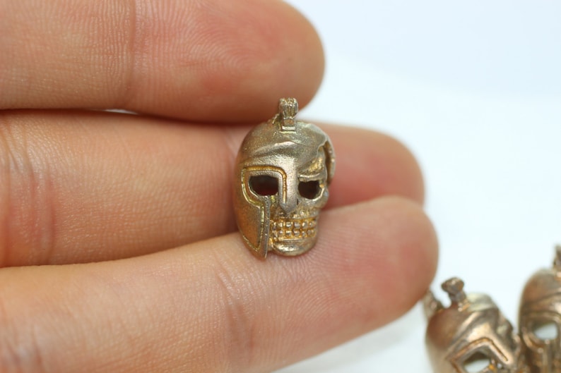 May include: A silver skull charm with a gold helmet design. The skull has a detailed face with eye sockets and teeth.