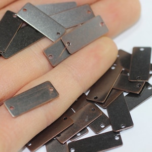 May include: A pile of small, rectangular metal tags with two holes in each. The tags are a mix of dark brown and silver colors.