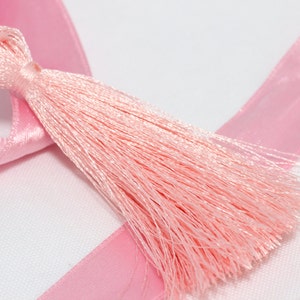 Può includere: Un nastro di raso rosa con un fiocco rosa attaccato. Il nastro e il fiocco sono su uno sfondo bianco.