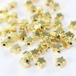 May include: A pile of gold-colored star-shaped beads, some with a hole in the center, on a white surface.