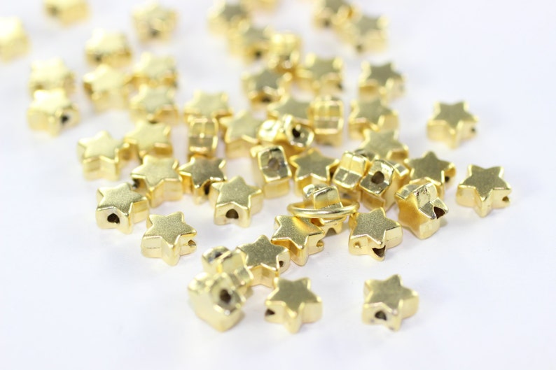 May include: A pile of gold-colored star-shaped beads, some with a hole in the center, on a white surface.