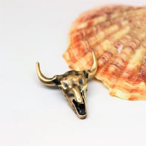 May include: A gold-colored bull skull pendant with a distressed finish. The pendant is sitting on a white surface next to a seashell.