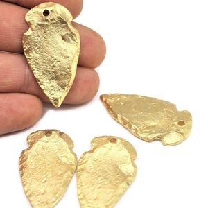 May include: Four gold-coloured arrowhead pendants with a hole at the top for stringing. The arrowheads have a rough, textured surface.