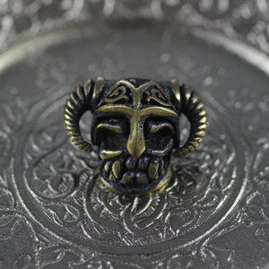 May include: A small, detailed, brass skull bead with horns and a helmet. The skull is black with gold accents. The bead is sitting on a silver tray with an ornate design.