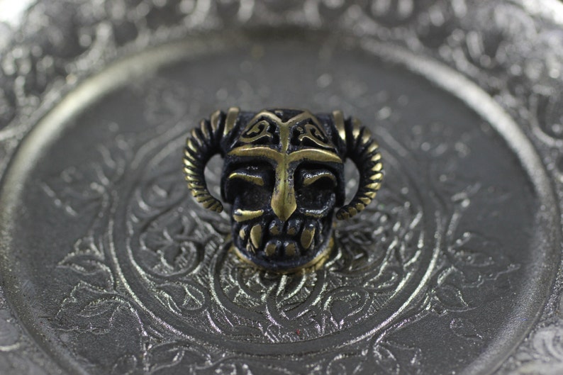 May include: A small, detailed, brass skull bead with horns and a helmet. The skull is black with gold accents. The bead is sitting on a silver tray with an ornate design.