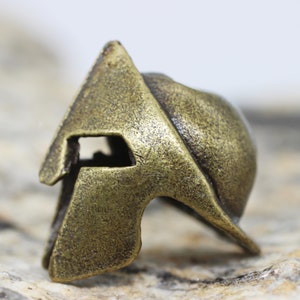 Op de afbeelding: Een bronzen Spartanen helmbedel met een gedetailleerd ontwerp. De helm heeft een verhoogde kam en een gezichtsmasker met een kleine opening voor de ogen.
