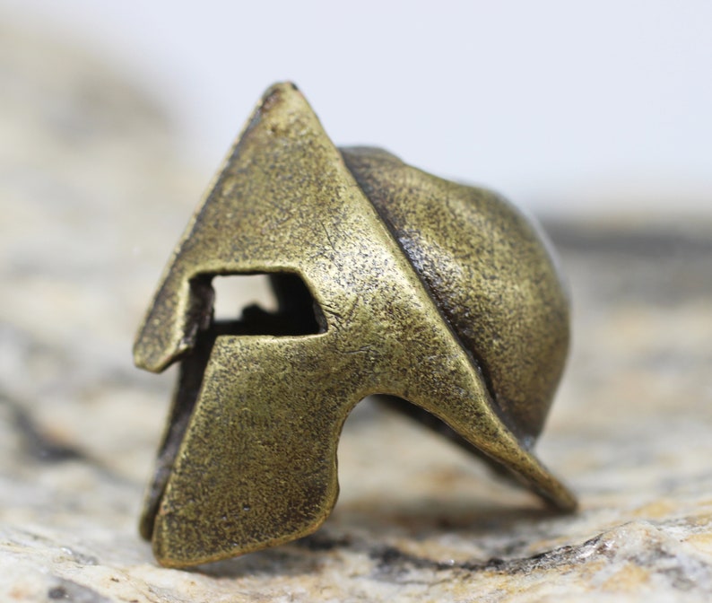 Op de afbeelding: Een bronzen Spartanen helmbedel met een gedetailleerd ontwerp. De helm heeft een verhoogde kam en een gezichtsmasker met een kleine opening voor de ogen.