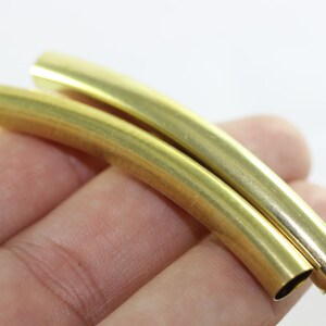 Puede incluir: Dos tubos de metal de color dorado, curvados en un ligero arco, se sostienen en una mano. Los tubos son lisos y brillantes.