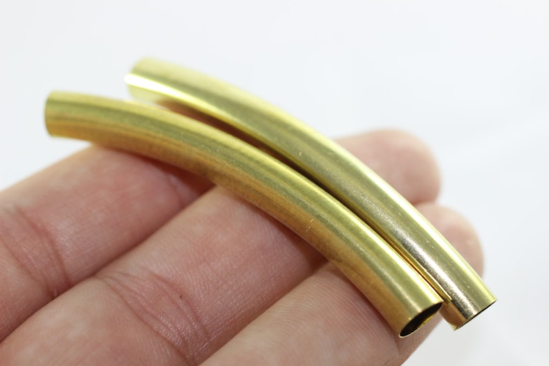 Puede incluir: Dos tubos de metal de color dorado, curvados en un ligero arco, se sostienen en una mano. Los tubos son lisos y brillantes.