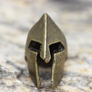 Op de afbeelding: Een gouden metalen Spartanen helm bedel. De helm heeft een puntige bovenkant en een gezichtsmasker met ooggaten.