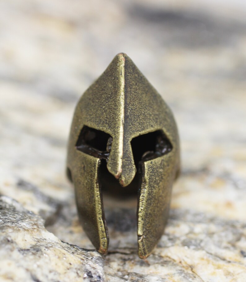 Op de afbeelding: Een gouden metalen Spartanen helm bedel. De helm heeft een puntige bovenkant en een gezichtsmasker met ooggaten.
