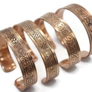 Puede incluir: Cuatro pulseras de cobre con intrincados diseños grabados. Las pulseras están apiladas una encima de la otra, con la pulsera más grande en la parte inferior y la más pequeña en la parte superior.