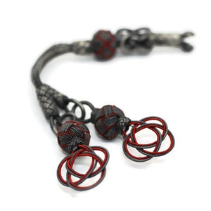 May include: A black and red knotted tassel with a silver chain. The tassel is made of braided cord and has a decorative knot at the end.
