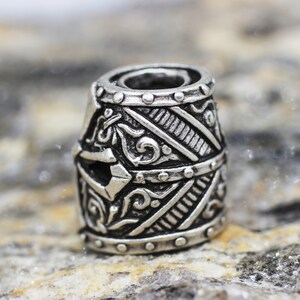 May include: A silver metal bead with an intricate design of a helmet. The bead has a detailed pattern of lines and swirls.