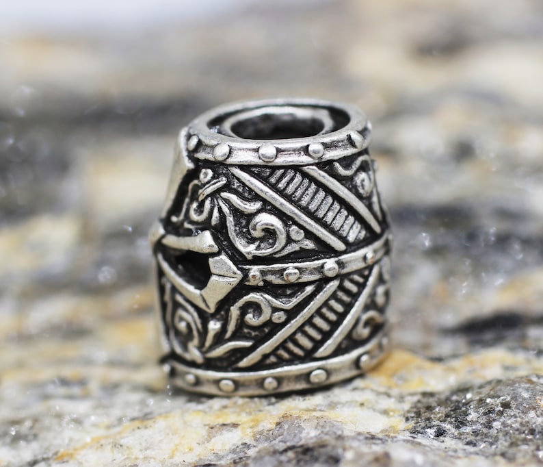 May include: A silver metal bead with an intricate design of a helmet. The bead has a detailed pattern of lines and swirls.