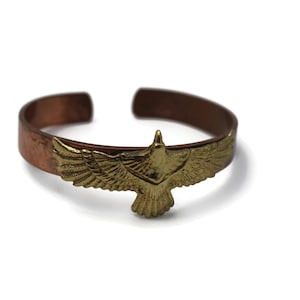May include: A copper-toned cuff bracelet with a brass eagle emblem. The open-ended bracelet features a detailed, outstretched eagle, adding a touch of nature-inspired style. The bracelet is designed to be worn on the wrist.