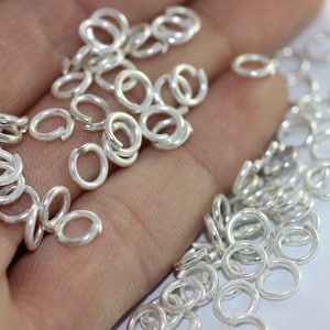 May include: A large quantity of silver jump rings, a type of jewelry finding, are scattered on a hand. The jump rings are small and circular, with a smooth, polished finish.