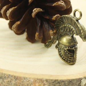 May include: A bronze-colored metal charm in the shape of a Roman helmet with a decorative crest. The charm is on a wooden surface.
